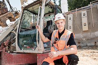 Foto eines Azubis, der vor einem Bagger kniet.