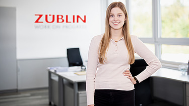 Foto von einer jungen Frau in einem Büro, die den Arm in die Seite stemmt und lächelt.
