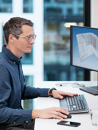 Bild von einem Mann, der an seinem Büroarbeitsplatz vor zwei Bildschirmen an einem Bauplan arbeitet.