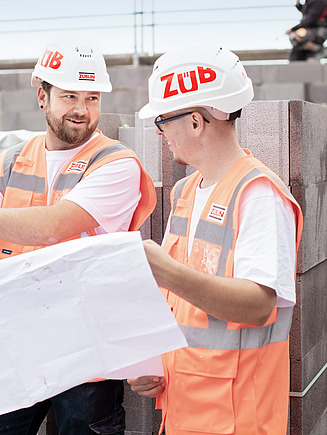 Zwei Kollegen stehen auf der Baustelle und haben einen Bauplan in der Hand