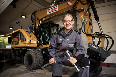 Bild einer Azubine, die vor einem Bagger sitzt mit Werkzeug in der Hand.