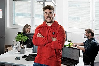 Junger Mann mit verschränkten Armen im Büro, lächelt in die Kamera. Im Hintergrund sind zwei weitere Personen am Schreibtisch sitzend.