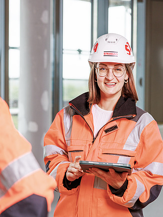 Eine junge Kollegin mit PSA und Tablet auf einer Baustelle.