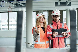 Zwei junge Menschen auf einer Baustelle, die sich unterhalten und die Baustelle anschauen.