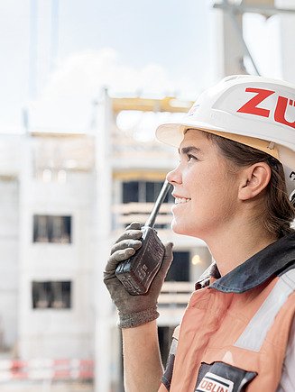 Junge Frau mit Bauhelm auf einer Baustelle, hält ein Funkgerät in der Hand.
