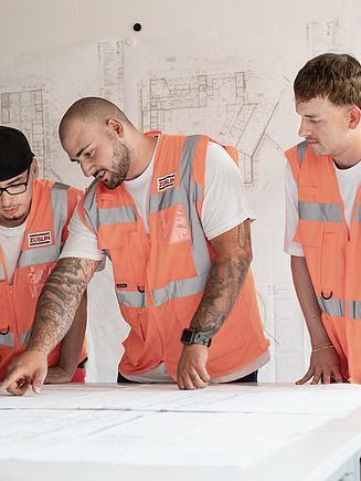Drei Kollegen stehen vor einem Tisch, der voller Baupläne ist