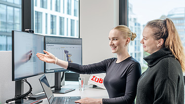 Foto von zwei Kolleginnen, die an einem Schreibtisch stehen und in den Bildschirm schauen.