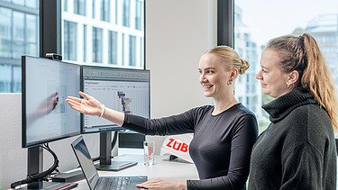 Foto von zwei Kolleginnen, die an einem Schreibtisch stehen und in den Bildschirm schauen. besprechen.