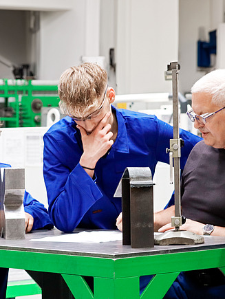 Mehrere Auszubildende stehen vor einer Werkbank. Ein Ausbilder erklärt etwas.