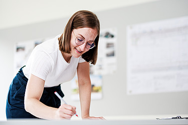 Eine Frau steht an einem Schreibtisch und hat einen Stift in der Hand.