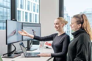 Foto von zwei Kolleginnen, die an einem Schreibtisch stehen und in den Bildschirm schauen. besprechen.
