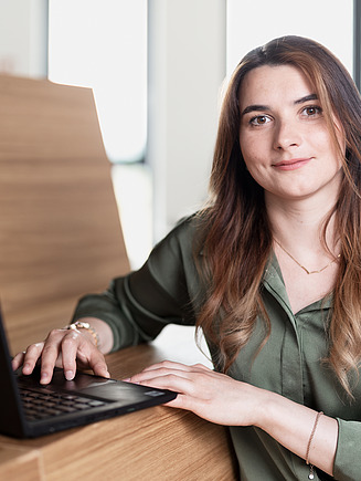 Foto einer Mitarbeiterin am Laptop, die in die Kamera schaut