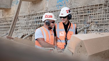 Bild eines Ausbilders und Azubis mit Schutzausrüstung auf einer Baustelle.